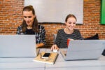 Two people sit at a table with laptops, smiling, in a room with a brick wall. Notebooks and a smartphone are on the table.