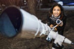 A person smiling while looking through a telescope outdoors at night, suggesting an interest in stargazing or astronomy.