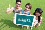 Three people standing on grass, one giving a thumbs up, holding a tablet displaying the word "WINNERS!"