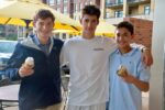 Three people standing together, smiling, with two holding ice cream cones. They are outdoors under yellow umbrellas, with buildings in the background.