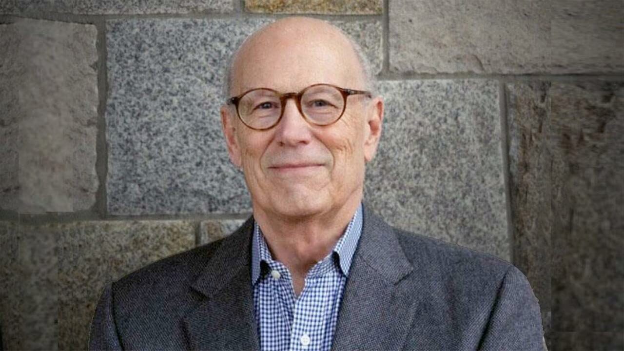 Headshot of a person with glasses, wearing a blazer and checkered shirt, standing in front of a stone wall.