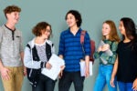 A group of five teenagers standing together, smiling and holding books and papers, suggesting a school or academic setting.