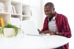 A person wearing a red and blue checkered shirt works on a laptop at a desk. Headphones rest around their neck, and there''s a plant and bookshelf in the background.