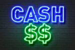 Neon sign displaying the word "CASH" in blue and two dollar signs "$$" in green against a brick wall.