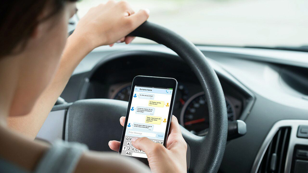 A person is texting on a smartphone while driving, showing a conversation on the screen. This represents distracted driving.