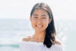 A person smiling at the camera with long dark hair, wearing a white off-shoulder top, standing by the ocean.