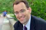 Headshot of a person wearing glasses and a suit, smiling in an outdoor setting with greenery in the background.