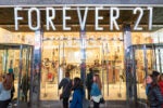 The image shows the entrance of a Forever 21 clothing store with people entering and passing by. The store is well-lit, displaying its interior with clothing and accessories.