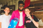 Three people are posing together inside a sneaker store. The person in the center is holding a shoe box and wearing sunglasses and a pink jacket. Sneakers are displayed on shelves in the background.