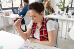 Person in a casual outfit, looking frustrated while holding a pencil to their forehead, seated at a desk with papers depicting mobile app designs, in a modern office.