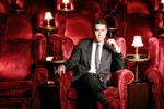 A person in a suit sitting in a plush red theater seat with similar empty seats and small lamps in the background, creating an elegant ambiance.