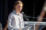 A person speaking at a clear podium with a microphone, wearing a striped shirt, against a dark background.