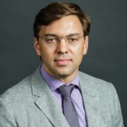 A headshot of a person wearing glasses, a gray suit, a purple shirt, and a patterned tie, against a dark background.