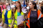 A group of people at a protest or march, with three individuals in the foreground holding megaphones and wearing bright safety vests. Signs and a crowd are visible in the background.