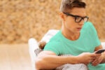 A person wearing glasses and a light blue shirt is seated on a couch, focused on using a smartphone. The background is a textured, warm-toned wall.