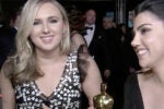 Two individuals dressed formally are smiling, one holding an Oscar-like statue, suggesting a red carpet or award event setting.