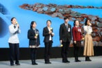 Six people in formal attire are holding yellow microphones, standing on a stage in front of a screen with coins displayed on it.