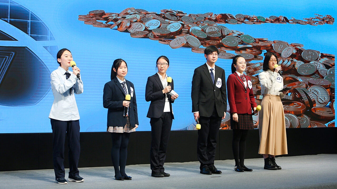 Six people in formal attire are holding yellow microphones, standing on a stage in front of a screen with coins displayed on it.