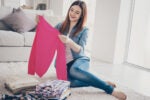 A person sitting on a floor at home, holding a pink sweater, smiling, with neatly folded clothes next to them.