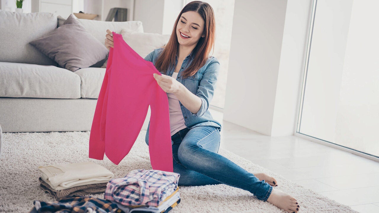 A person sitting on a floor at home, holding a pink sweater, smiling, with neatly folded clothes next to them.