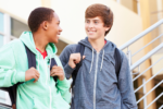 Two teenagers wearing hoodies and backpacks stand outside a building, smiling and talking.