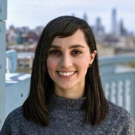 Headshot of a person with shoulder-length dark hair, smiling, wearing a gray sweater. The background is blurred with a cityscape view.