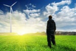 A person is standing in a green field, gazing at a wind turbine against a cloudy sky with sunlight in the background. It represents renewable energy and environmental awareness.