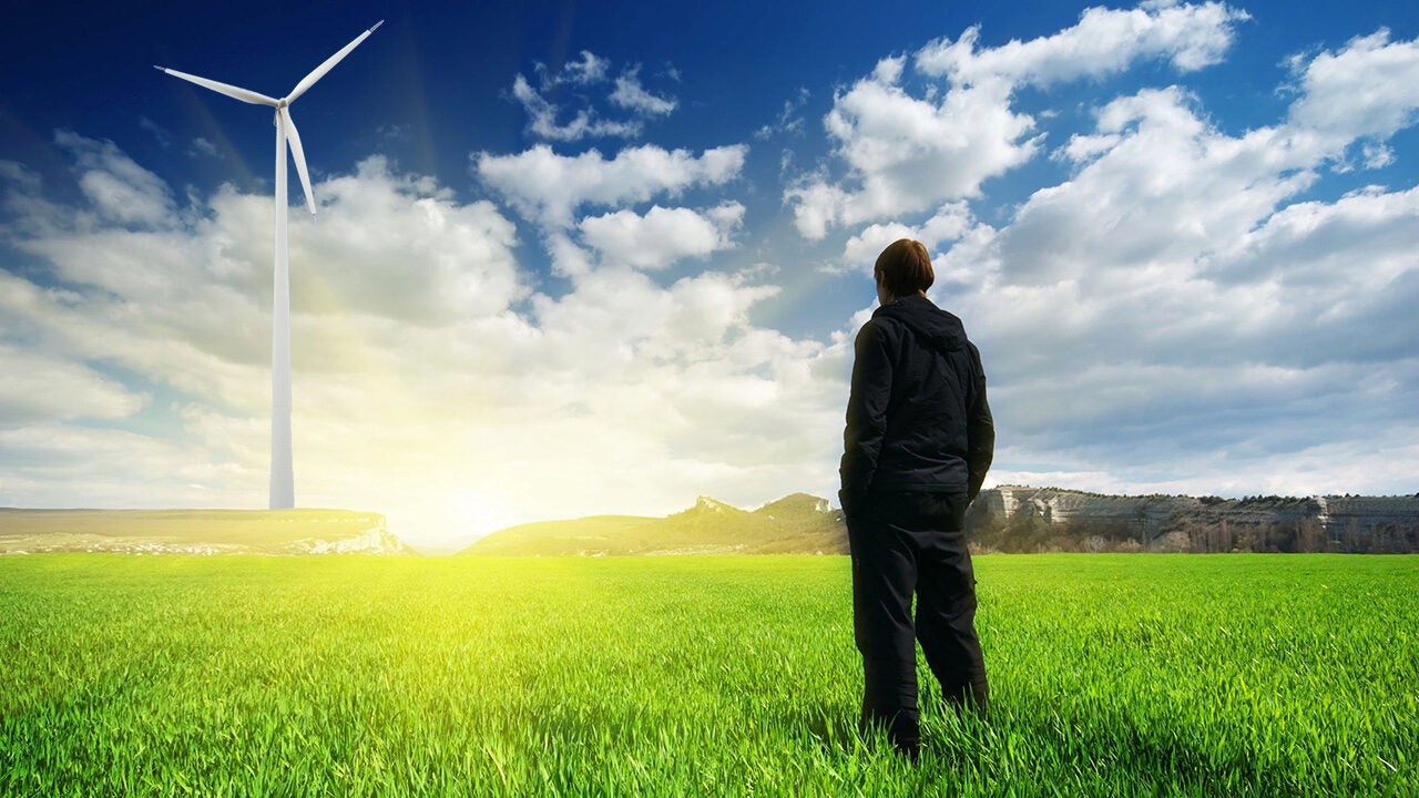 A person is standing in a green field, gazing at a wind turbine against a cloudy sky with sunlight in the background. It represents renewable energy and environmental awareness.