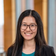 A headshot of a person with long, dark hair and glasses, smiling, wearing a black jacket and teal top, with a blurred background.