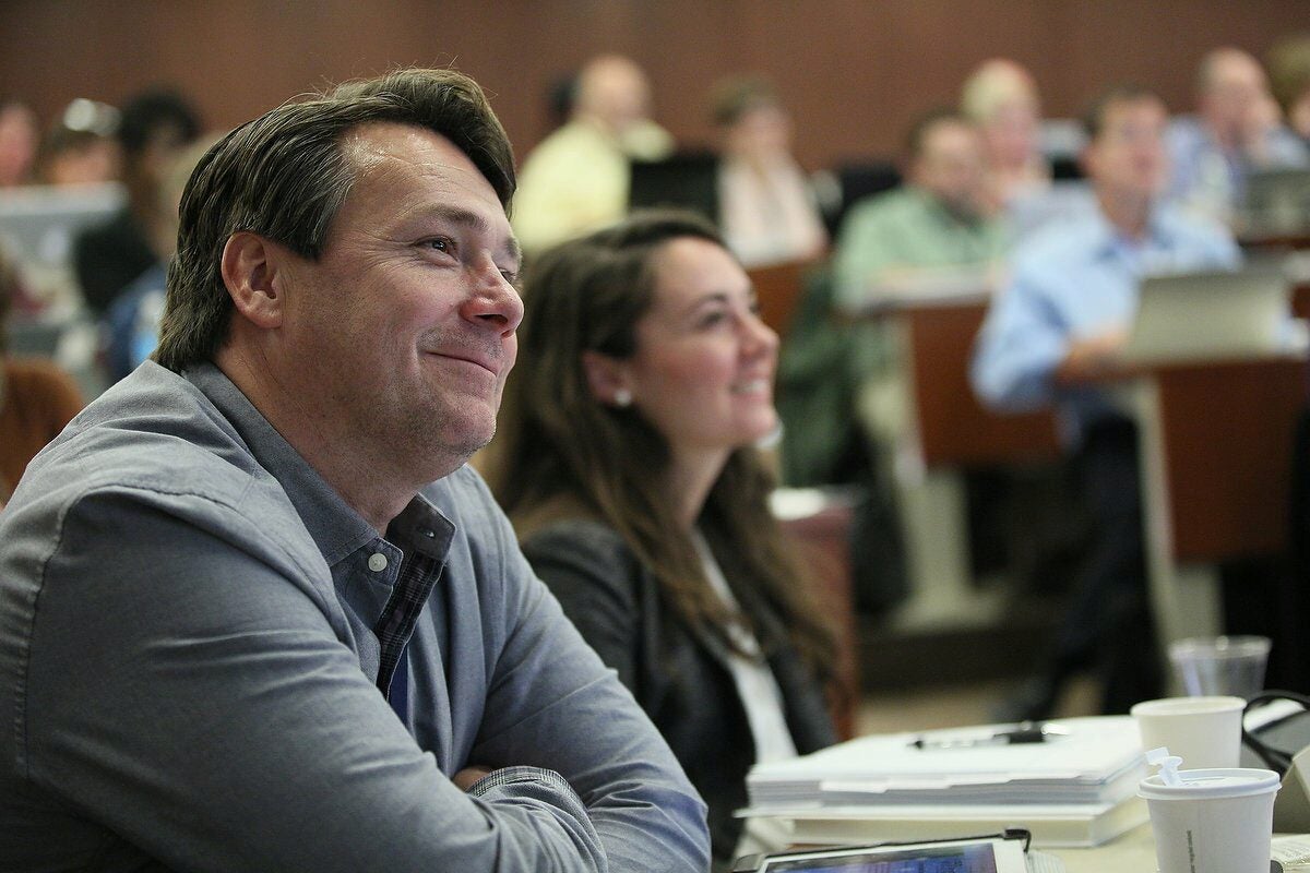 A classroom scene with people attentively listening, featuring a smiling individual in focus. Notebooks and cups are on the table.