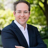 A person wearing glasses and a dark suit jacket, smiling with arms crossed, stands outdoors with trees and greenery in the background.