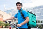 A person wearing a helmet and a blue shirt is riding a bicycle while carrying a large delivery backpack. The scene is set in an urban area with modern buildings in the background.