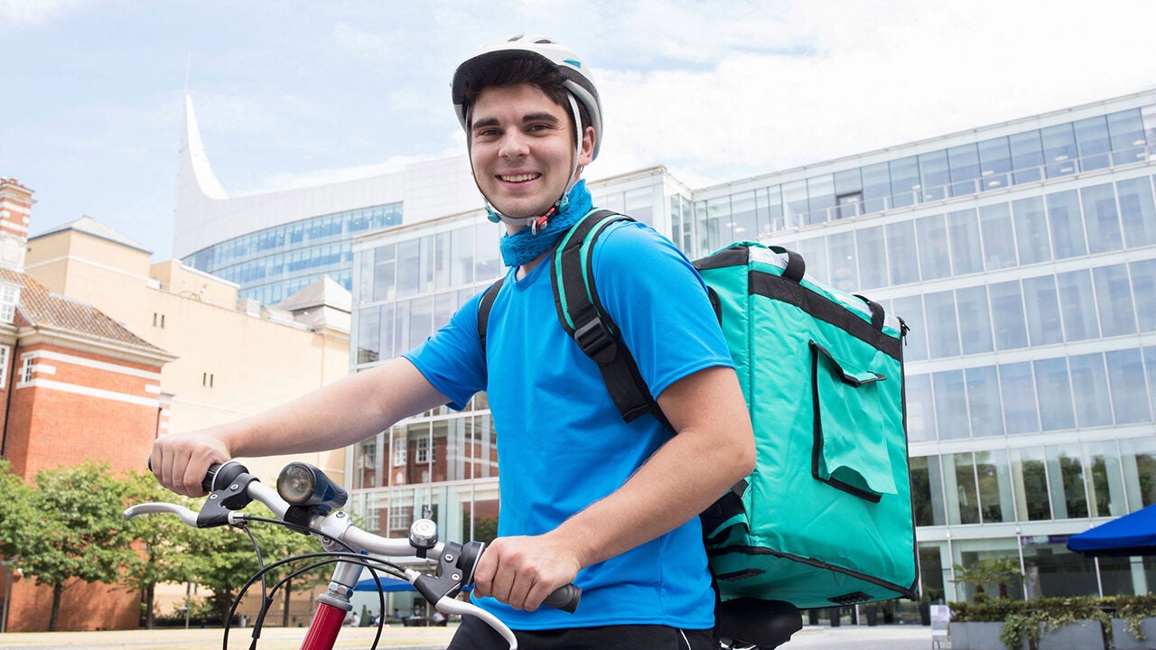 A person wearing a helmet and a blue shirt is riding a bicycle while carrying a large delivery backpack. The scene is set in an urban area with modern buildings in the background.