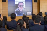 An audience watches a video call presentation on a large screen. The person on the screen wears sunglasses and a polo shirt. Attendees hold up phones to record the event.