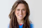 Headshot of a smiling person with long brown hair, wearing a blue and red shirt.
