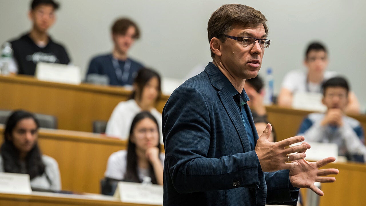 A person in a suit is speaking in front of a group of seated individuals, likely in a classroom or lecture setting. The audience is focused on the speaker.