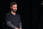 A person with a beard, wearing a dark shirt, sits against a black background with a Wharton logo in the top right corner.