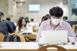 A person wearing a face mask is focused on a laptop in a library or study area. Several other people are seated nearby, engaged in their own activities.