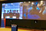 A conference room displays a large screen with two separate images. The left image shows a group of people in suits, possibly presenting. The right image is a scene from a film or digital artwork. A Wharton School banner and podium are visible in the