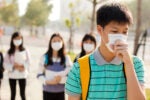 A group of young people wearing face masks outdoors. One person in the foreground is coughing and appears concerned or unwell. It represents health precautions and awareness.