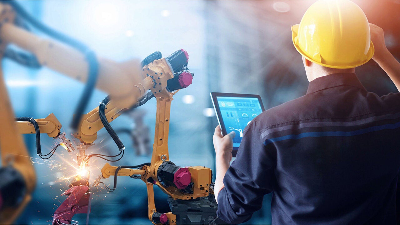 An engineer wearing a yellow hard hat operates a tablet while overseeing robotic arms welding metal, representing automation in manufacturing.