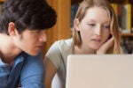 Two people are focused on a laptop screen in a library setting, suggesting a collaborative study session or research project.