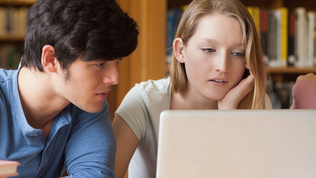 Two people are focused on a laptop screen in a library setting, suggesting a collaborative study session or research project.