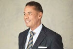 A person in a suit and tie, smiling and looking to the side, set against a plain background.