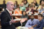 A person in a suit gives a lecture or presentation to an audience in a lecture hall or classroom setting. Audience members listen attentively.