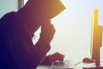 A person wearing a hoodie, sitting at a desk, using a computer. The image suggests themes of cybersecurity or hacking.