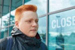 A person with short red hair wearing a dark jacket stands in front of a building with large windows. The word "CLOSED" is visible on the glass.