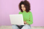 Person in a green sweater sitting cross-legged using a laptop against a light pink background.