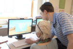 A younger person assists an older person with a computer in a classroom setting. The screen displays a map application.