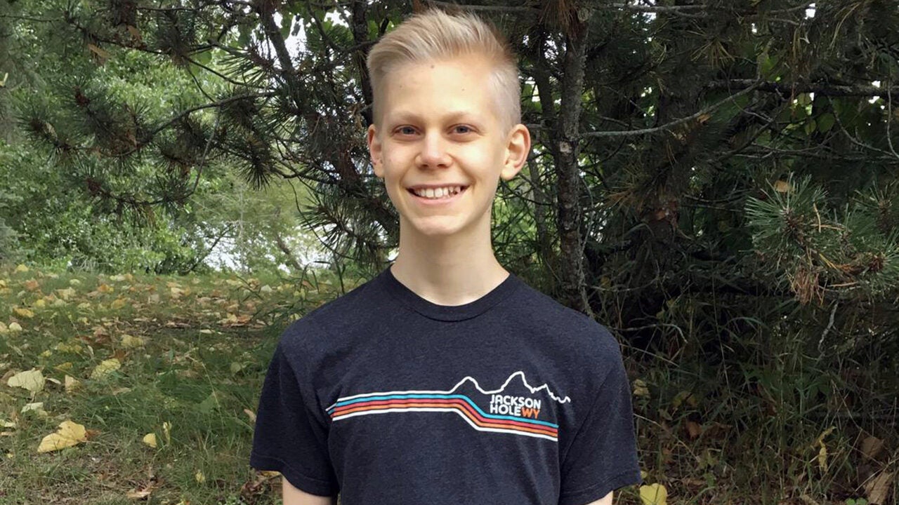 A smiling person with short blonde hair stands outdoors in front of a tree, wearing a t-shirt that reads "Jackson Hole WY." Lush greenery and some fallen leaves are visible.